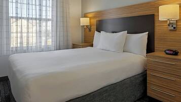 A hotel room with a large bed, wooden headboard, two bedside lamps, a nightstand, and a window with sheer curtains.