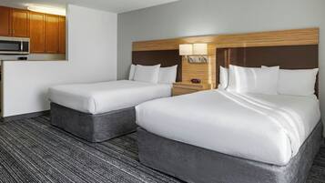 Quarto de hotel com duas camas, micro-ondas, pequeno espaço de cozinha e mesa de cabeceira com luminária.