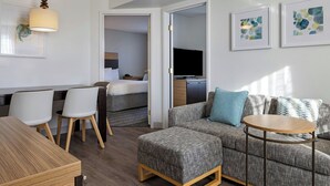 A modern hotel room with a wooden dining table, grey upholstered sofa, and a small round table.