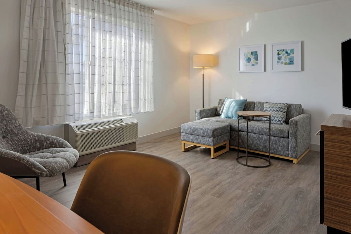 Ein modernes Wohnzimmer mit grauem Sofa, Hocker und Ohrensessel, einem kleinen runden Tisch und einer stehenden Stehlampe.