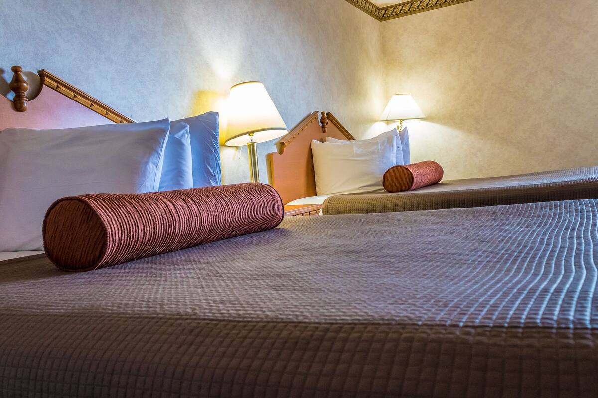 Ein Hotelzimmer mit zwei Betten, jeweils mit Kopfteil, Kissen und einer zusammengerollten Decke. Auf jedem Bett stehen zwei Nachttischlampen.