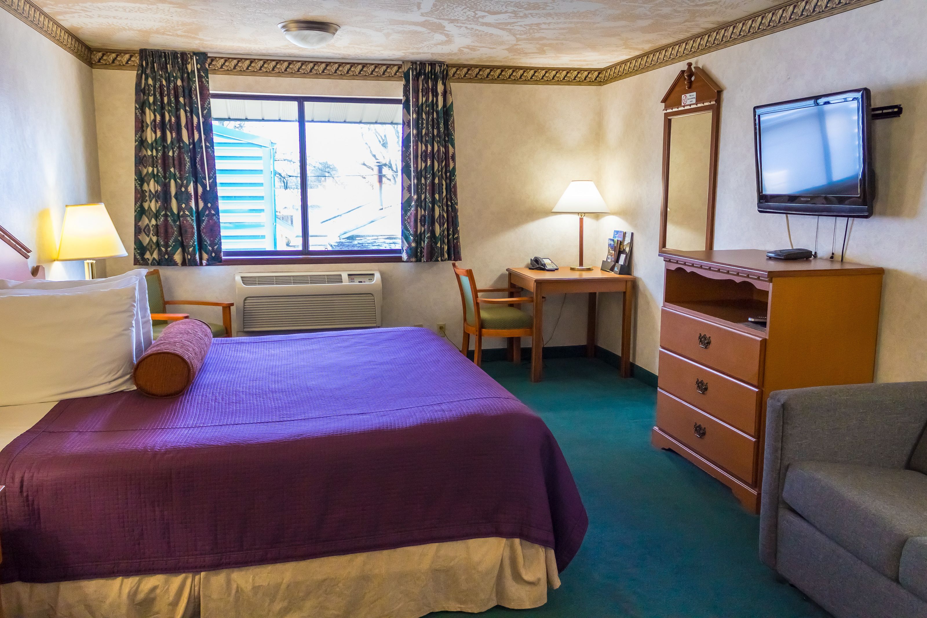 A hotel room with a bed, desk, chair, dresser, TV, and window with curtains.