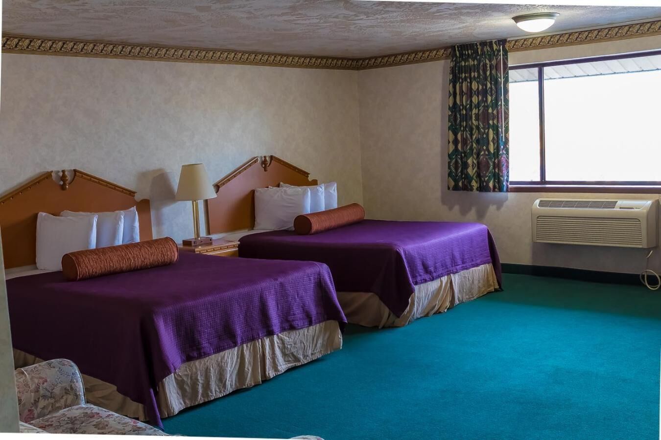 Habitación de hotel con dos camas, una ventana con cortinas, un equipo de aire acondicionado y una silla con estampado floral.
