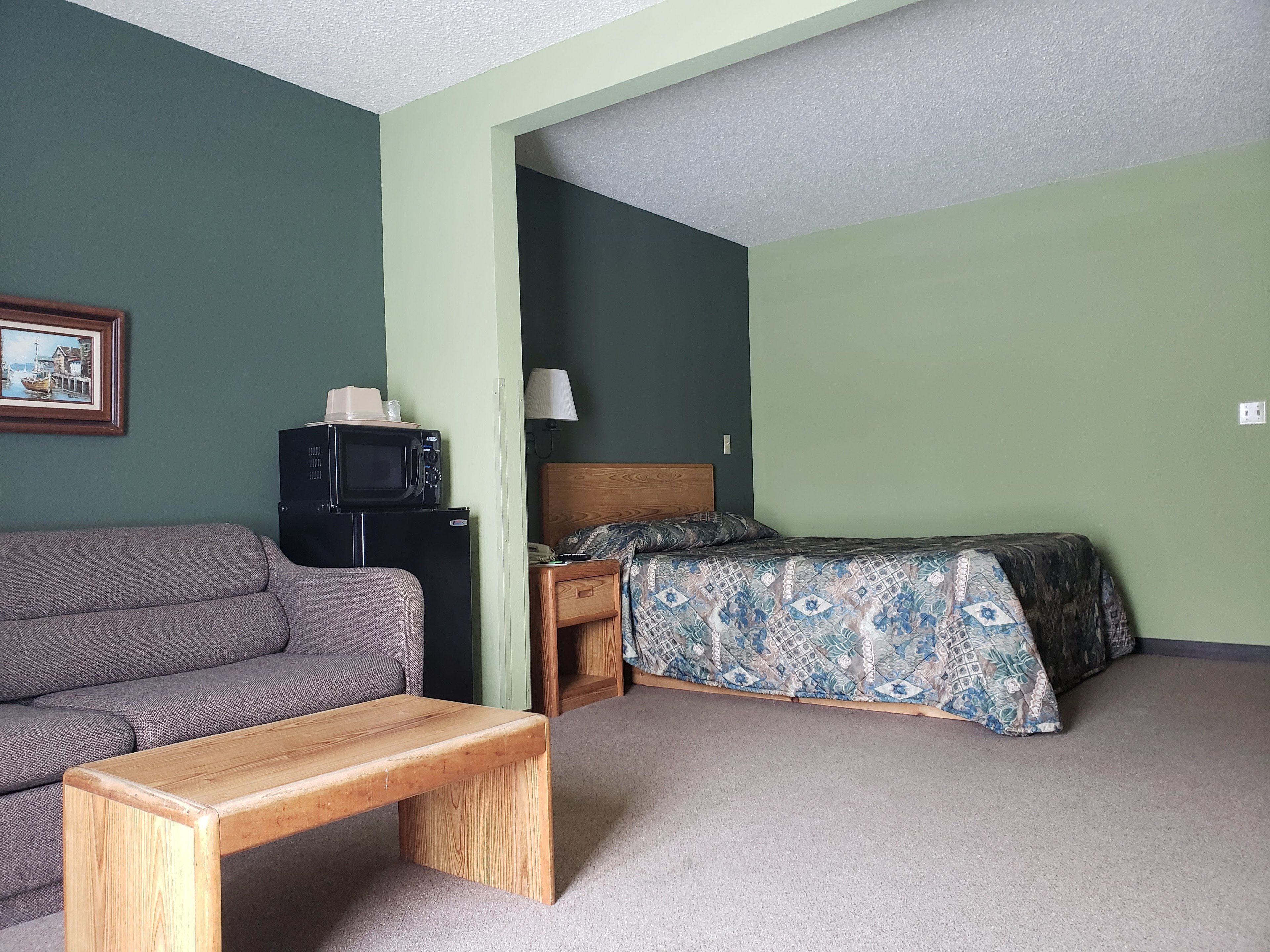 Een kamer met een bank, een houten zitbank, een bed met beddengoed met een patroon, een zwarte koelkast en een magnetron erboven.