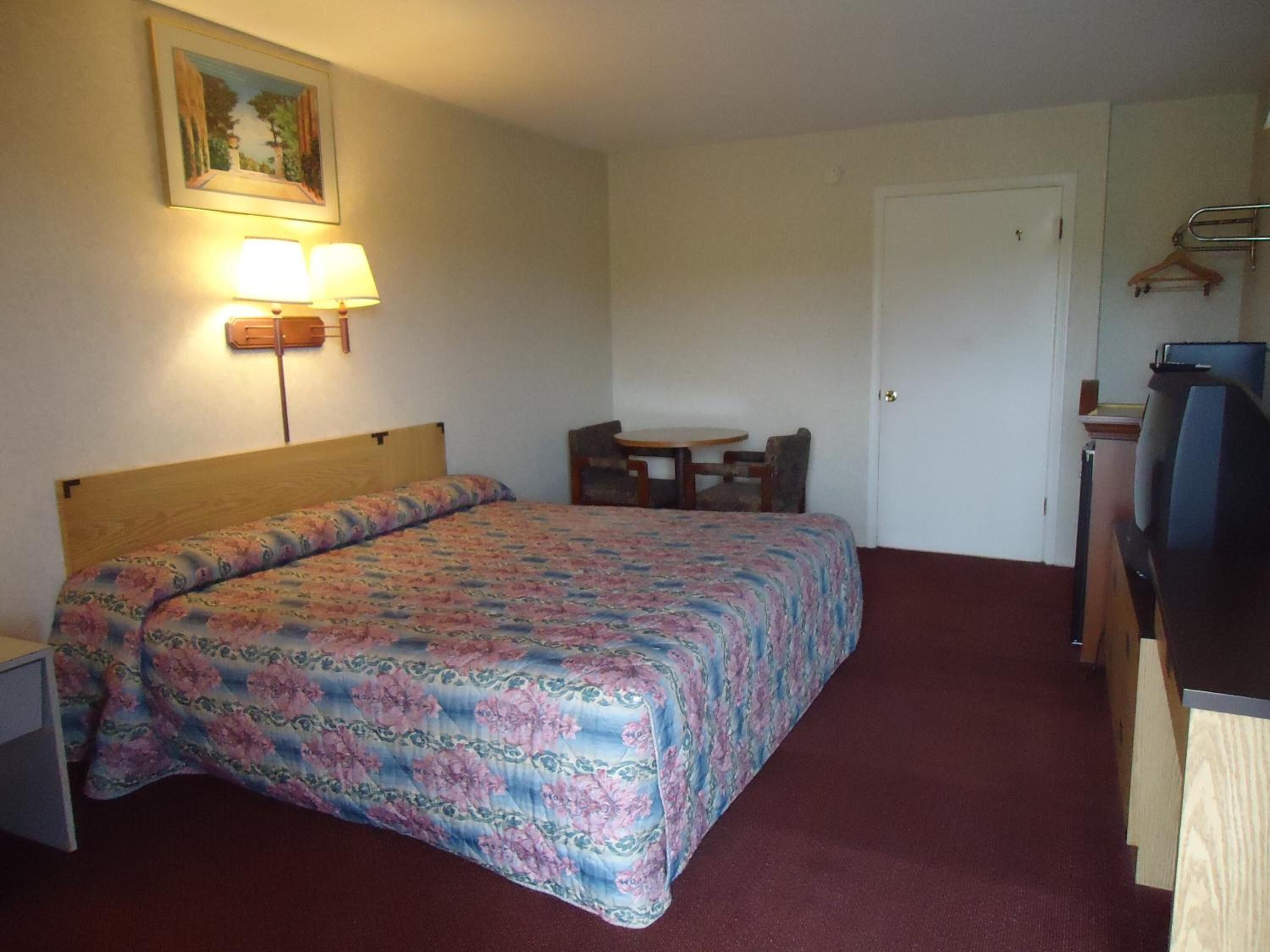 Una habitació d'hotel amb un llit, una taula de nit, una cadira, un televisor i un quadre penjat a la paret.