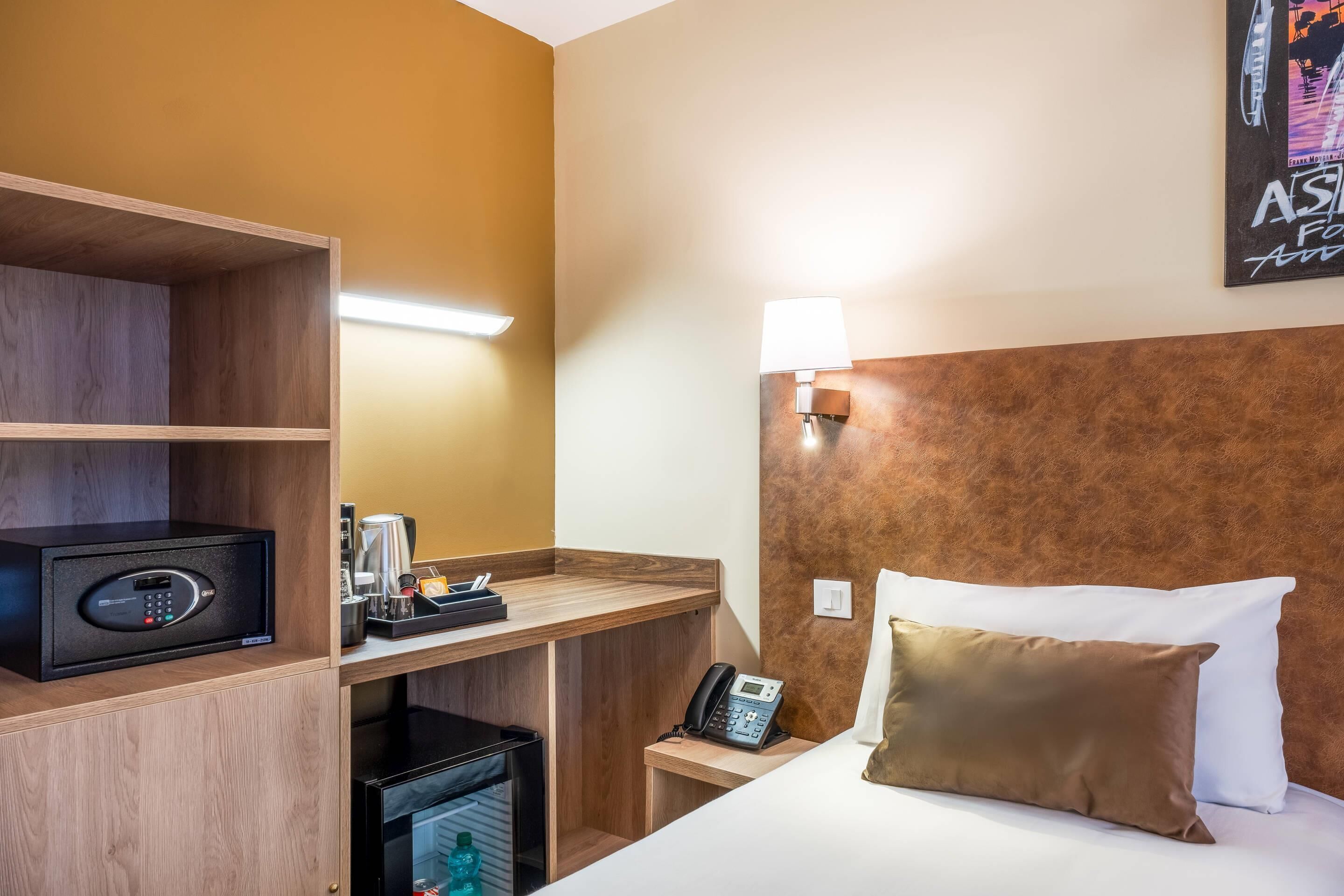 A hotel room with a bed, a desk with a telephone, a minibar, and a wall-mounted lamp.