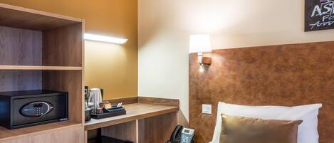 A hotel room with a bed, a desk with a telephone, a minibar, and a wall-mounted lamp.