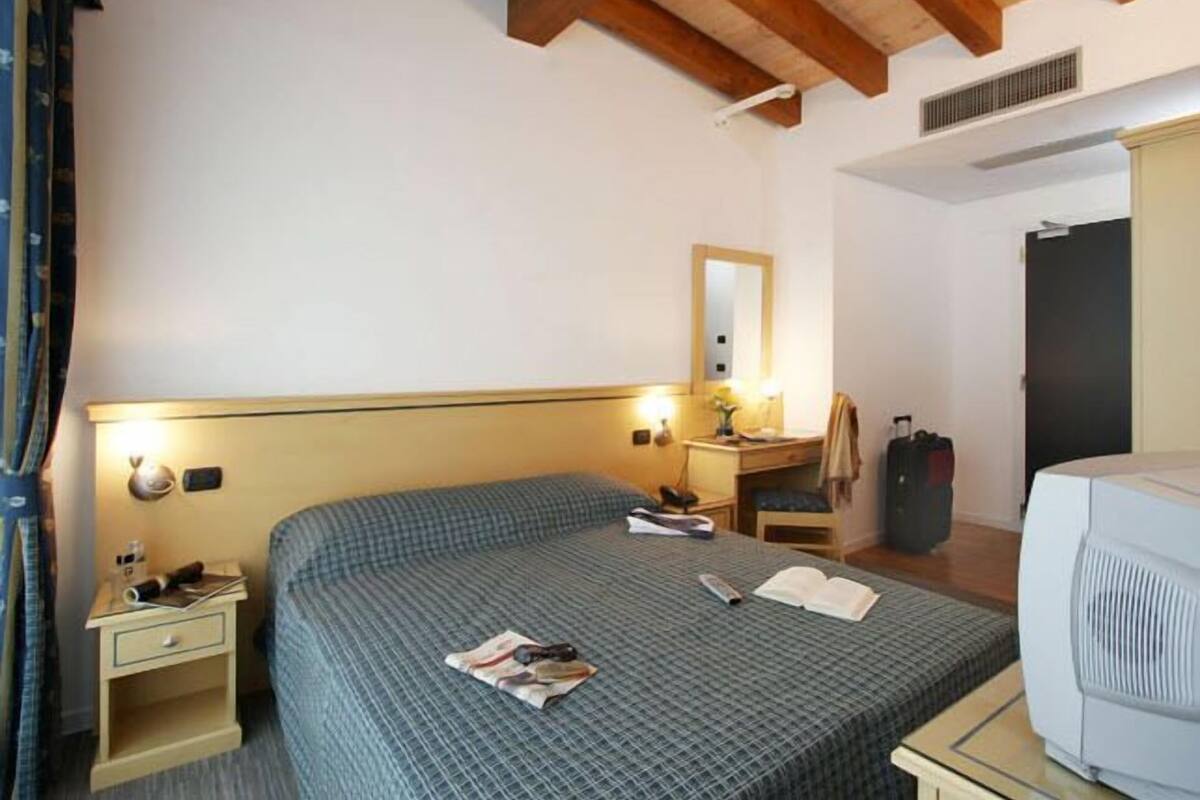 A hotel room with a bed, bedside tables, a desk with a lamp, a mirror, a wardrobe, and a television.