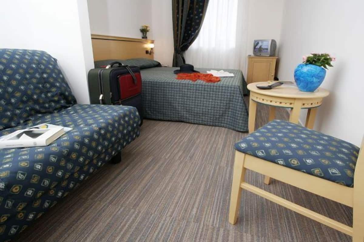 A hotel room with two beds, a small table, a chair, and a suitcase.