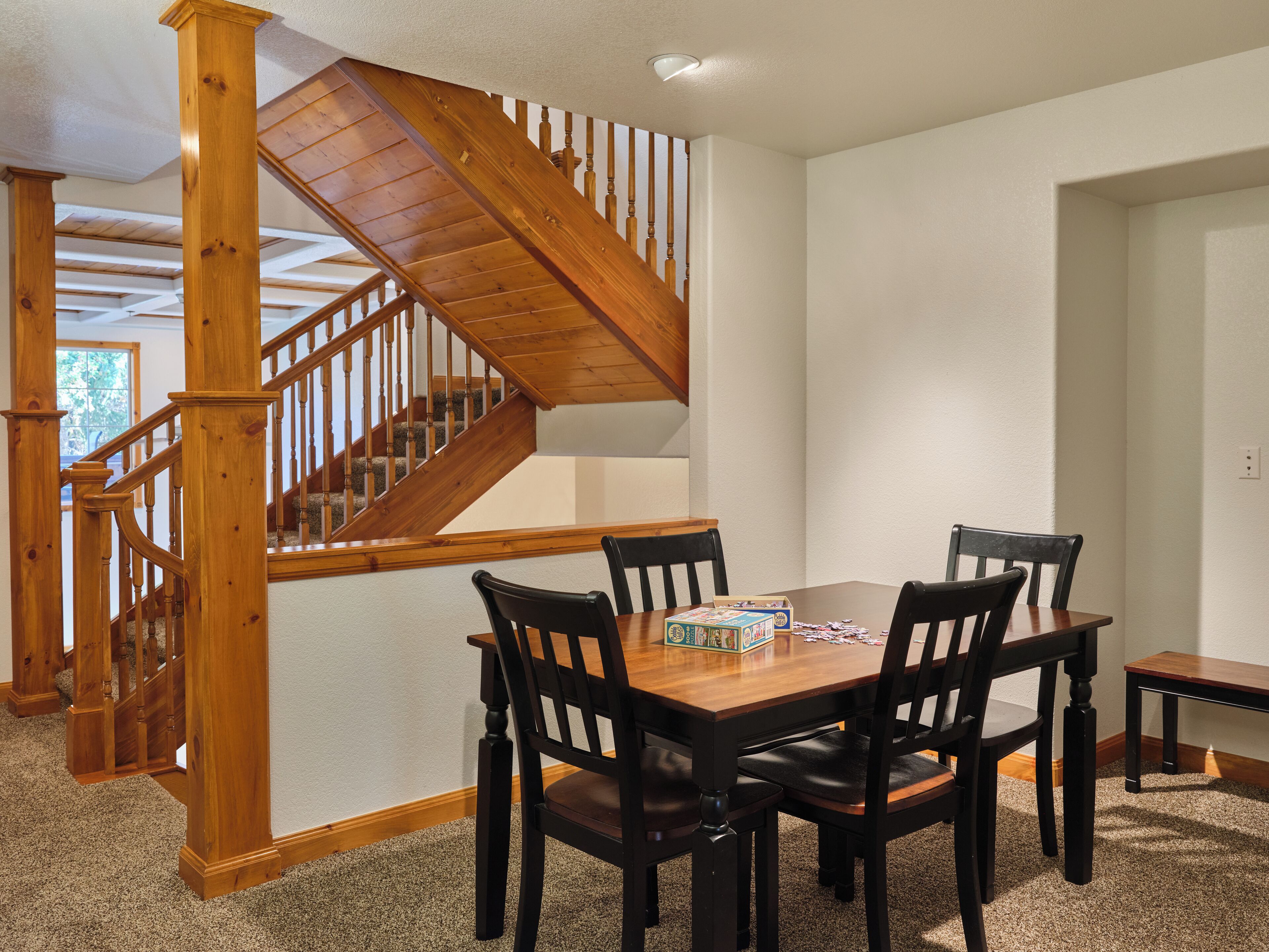A dining area with a wooden table and chairs, a staircase with wooden railings, and a carpeted floor.