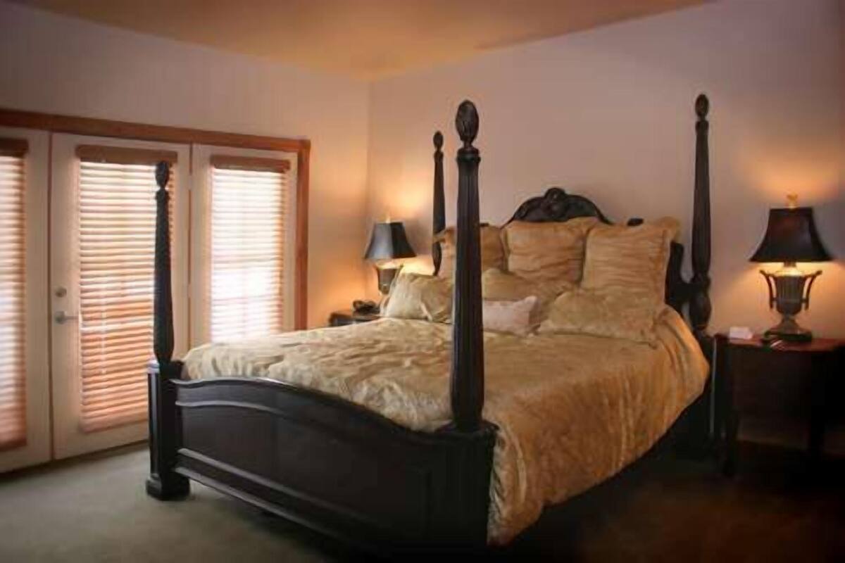 A bedroom with a four-poster bed, a nightstand with a lamp, and a door with blinds.