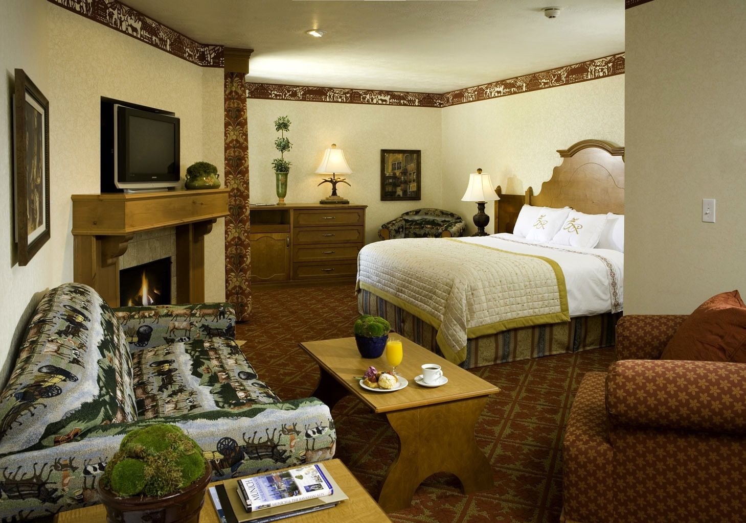 A hotel room with a bed, a sofa, a coffee table, a television, a fireplace, and a small table with a book and a glass of juice.
