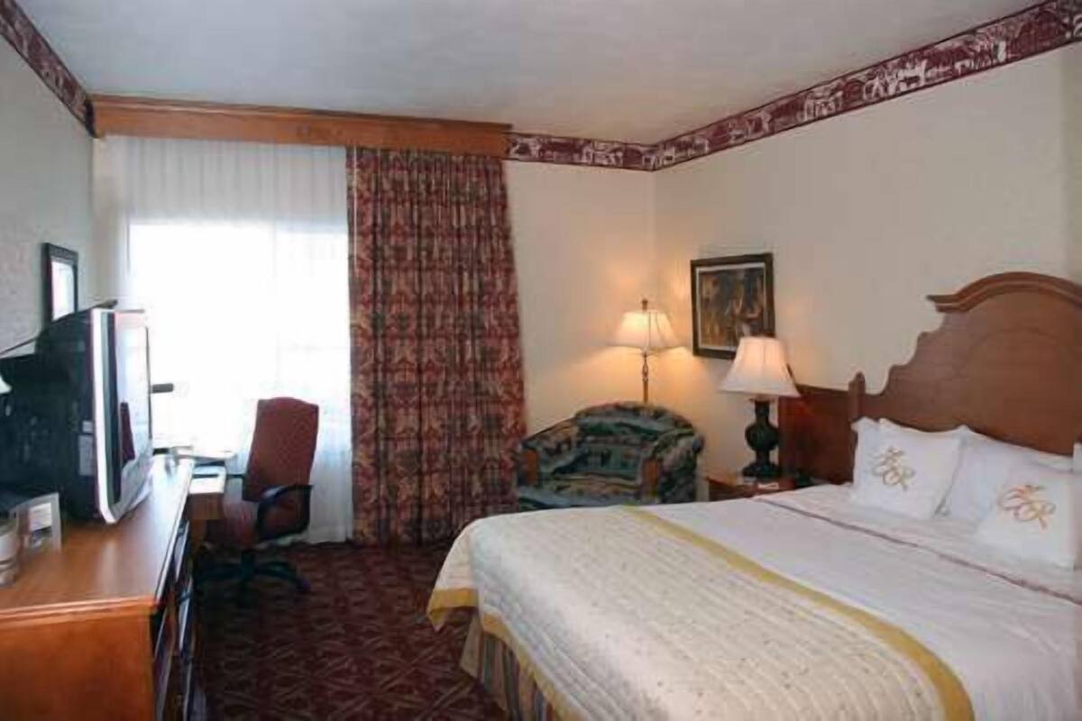 A hotel room with a bed, a desk with a television, a chair, a lamp, and a window with curtains.
