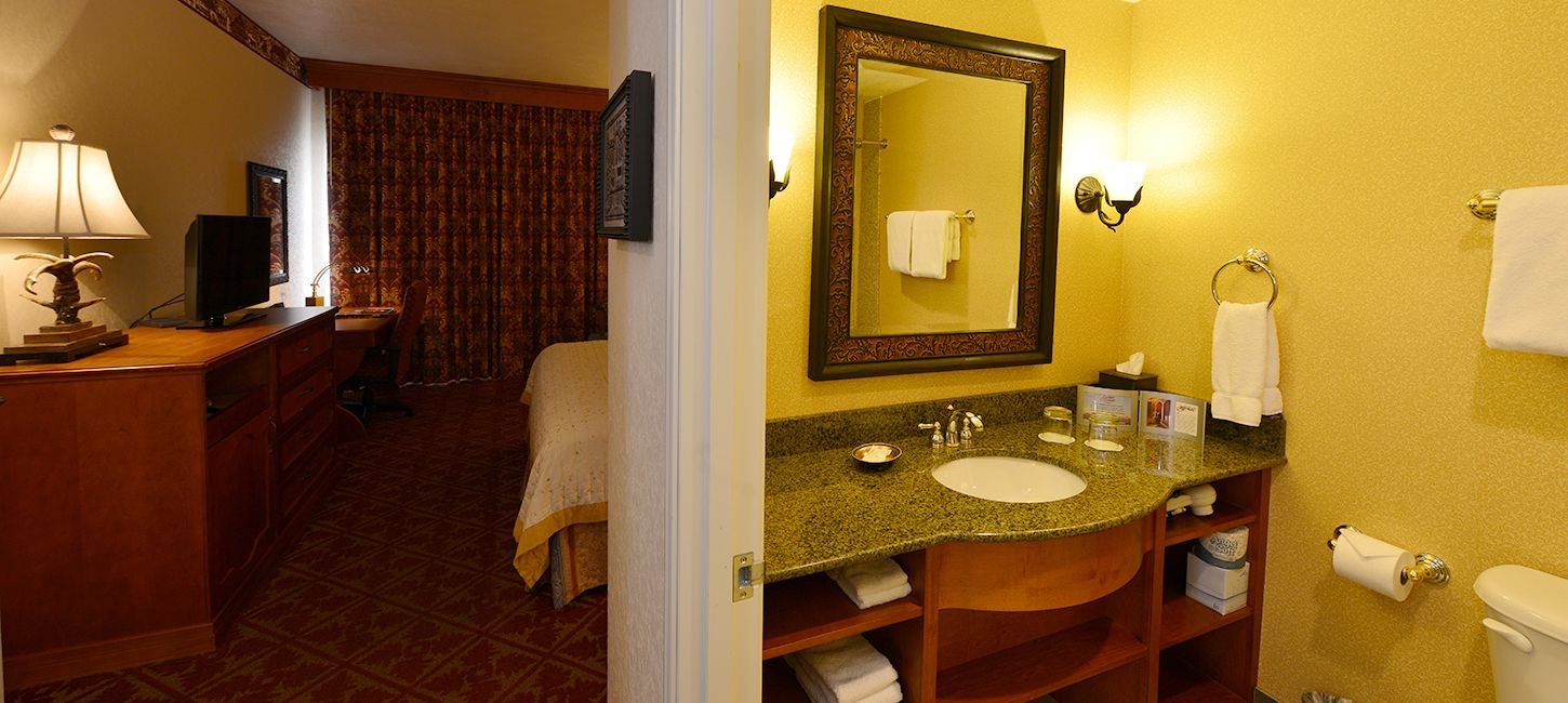 A hotel room with a wooden desk, a television, a bed with a beige bedspread, a bathroom with a granite countertop sink, a mirror, and a toilet.