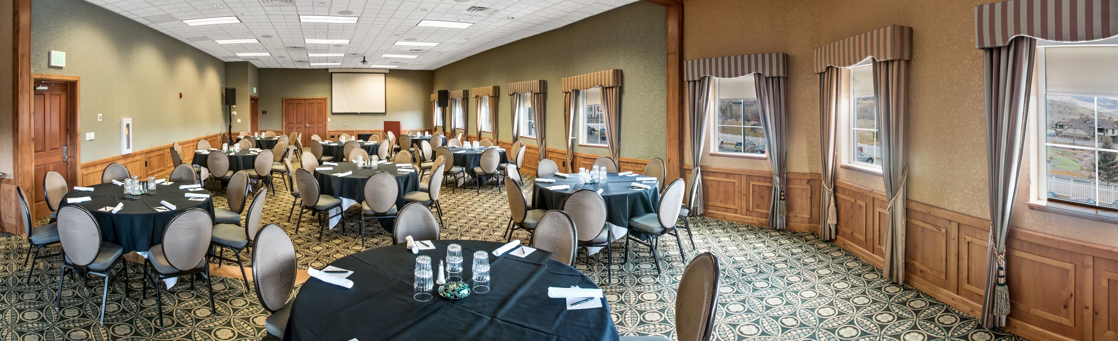 A conference room with multiple tables and chairs, large windows with curtains, and a carpeted floor.