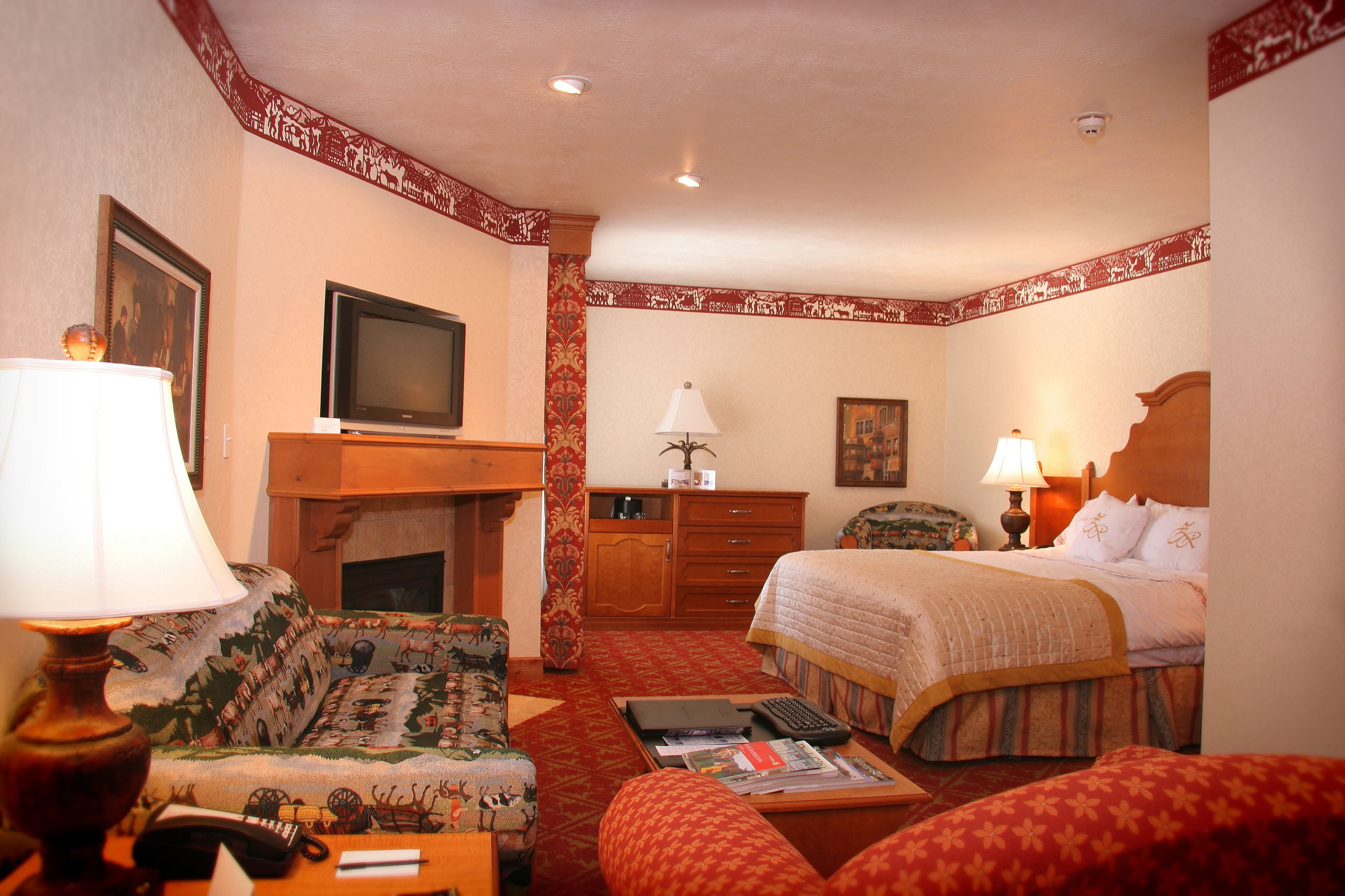 A hotel room with a bed, a sofa, a desk with a lamp, a television, and a fireplace.