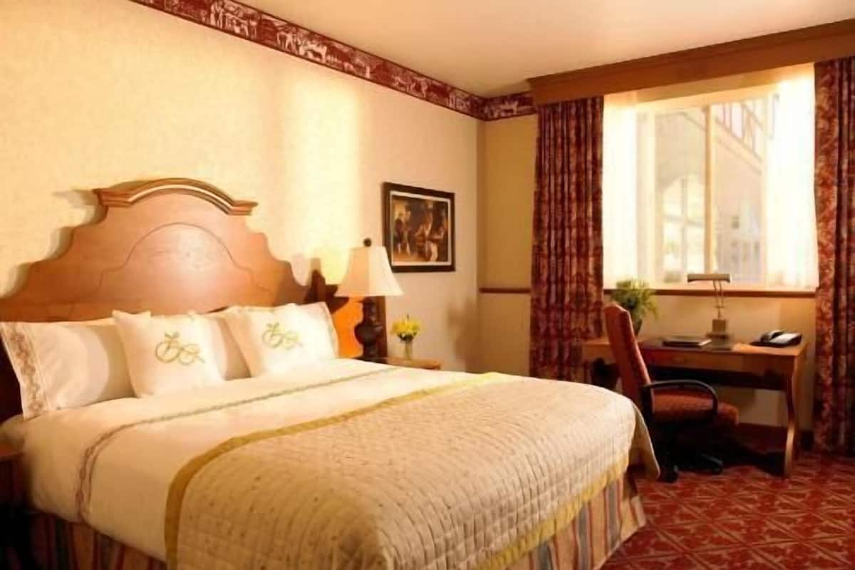 A hotel room with a bed, a desk, a chair, a lamp, a picture on the wall, and a window with curtains.