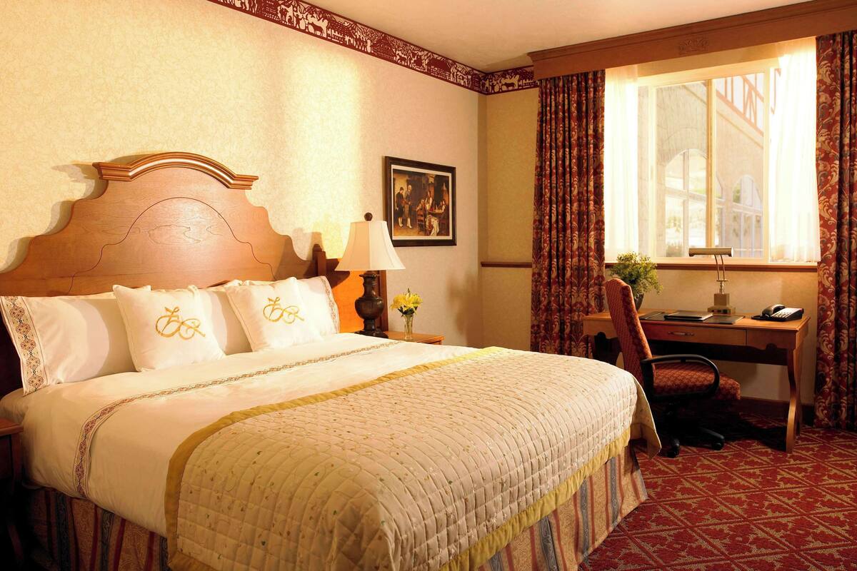 A hotel room with a large bed, a desk with a chair, a window with curtains, and a painting on the wall.
