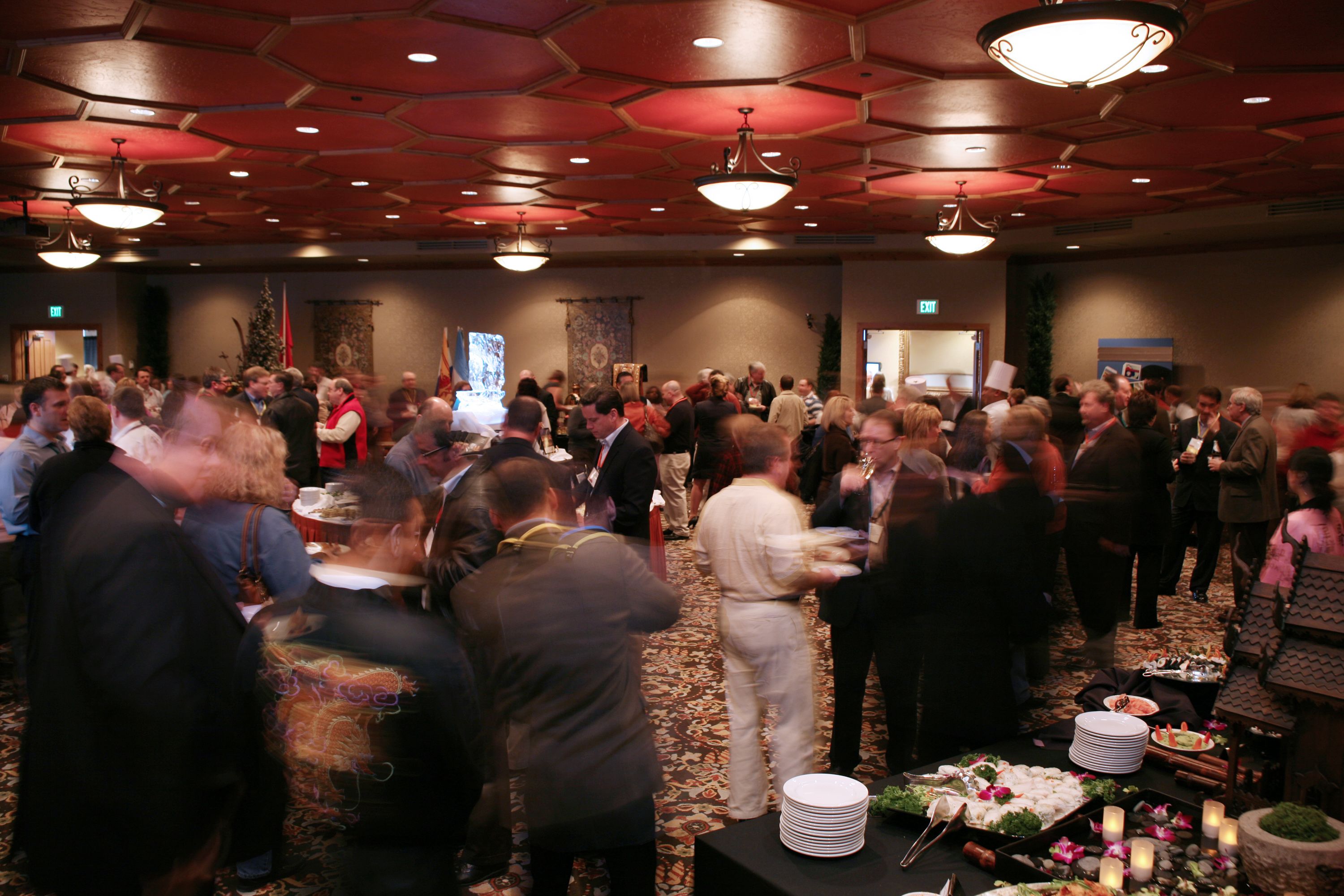 A crowded indoor event with tables and chairs, a patterned carpet, and a chandelier.