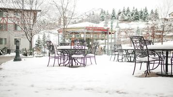Outdoor seating with snow-covered tables and chairs, a building with a red roof, and snow-covered trees in the background.