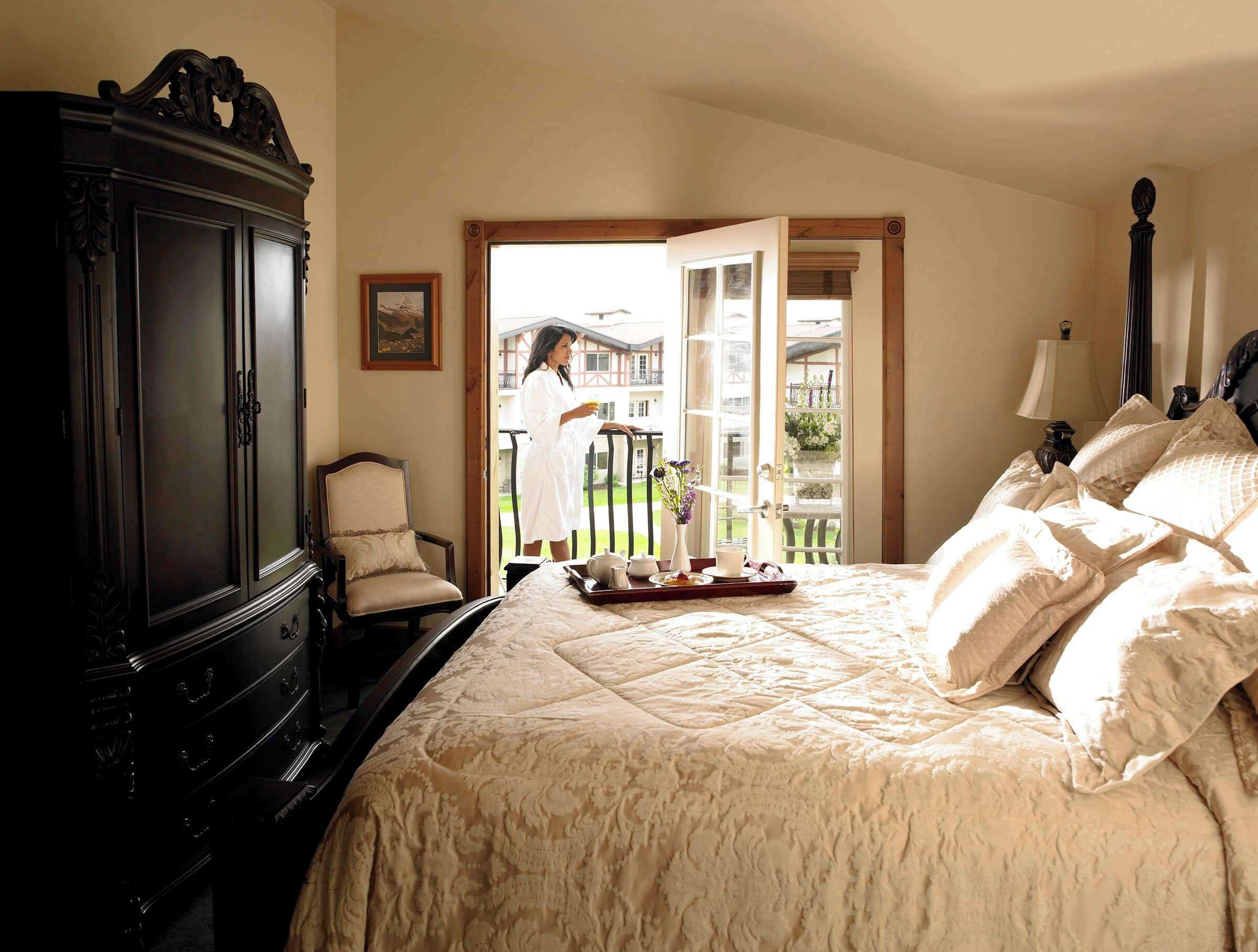 A bedroom with a large bed, a dark wooden wardrobe, a chair, and a balcony with a view.