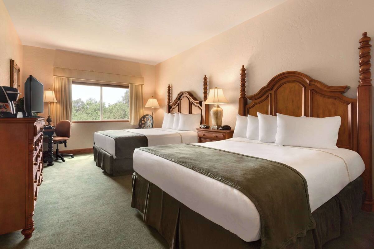 A hotel room with two beds, a desk, a television, and a window with a view of trees.