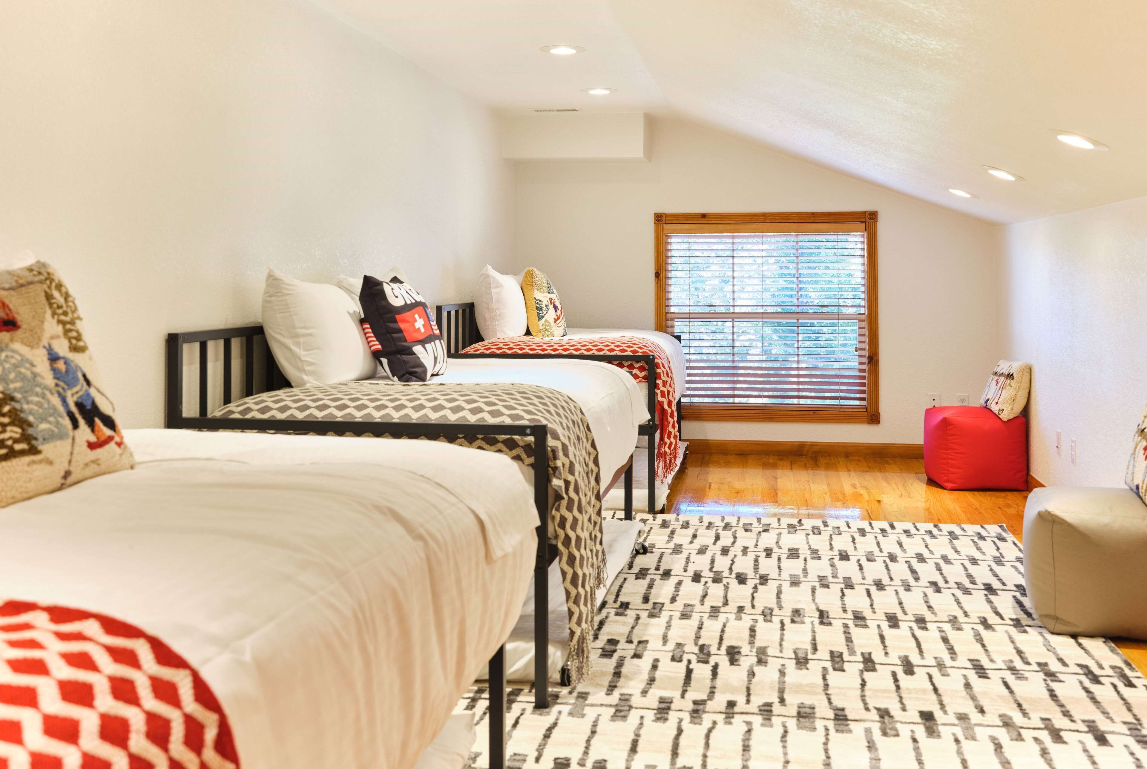 A bedroom with two beds, a window with blinds, a red ottoman, and a patterned rug.