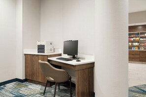 Ein Hotelzimmer mit Schreibtisch, Computer, Stuhl, Kaffeemaschine und einer Minibar im Hintergrund.
