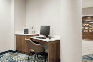 A hotel room with a desk, a computer, a chair, a coffee machine, and a snack bar in the background.