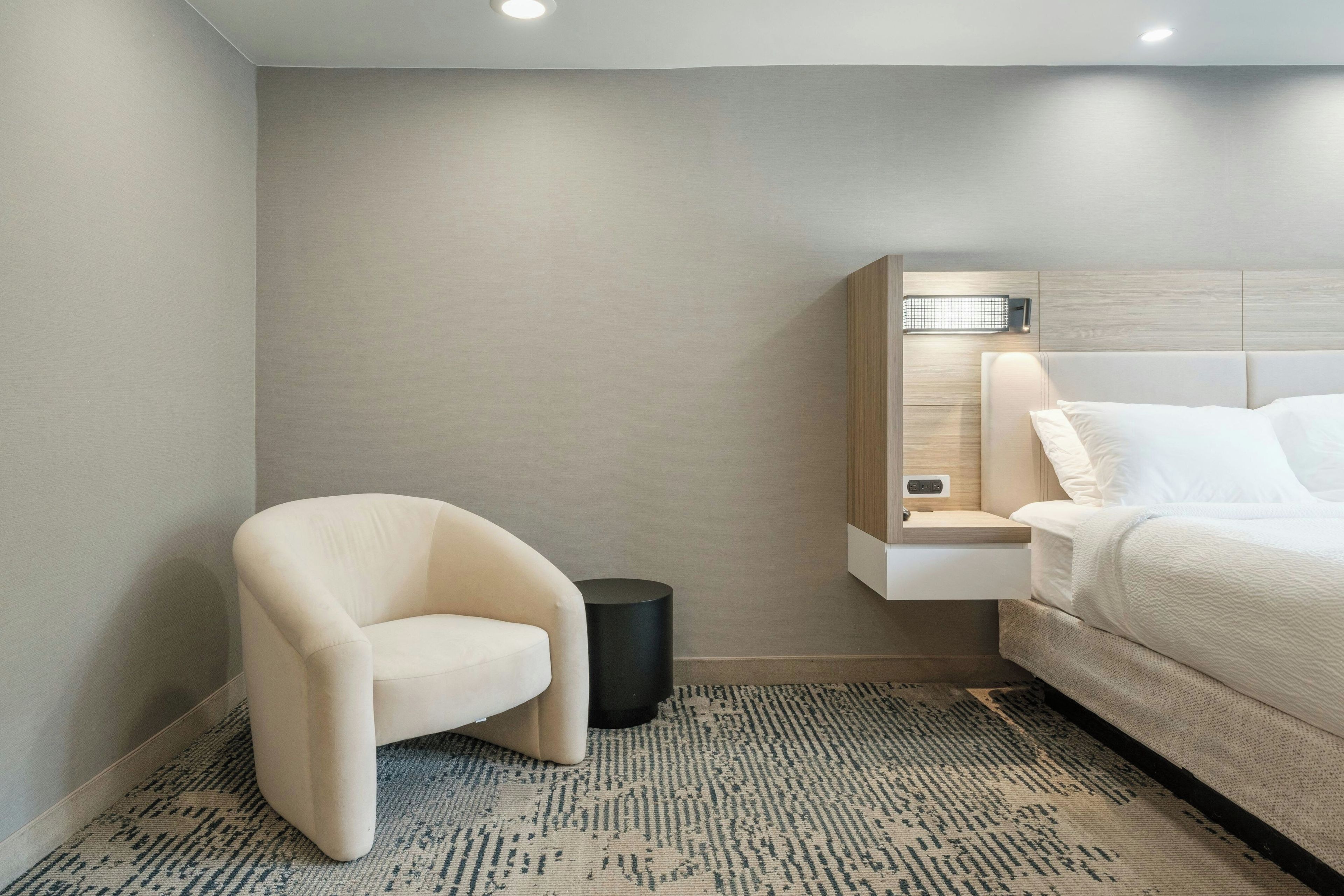 Moderni hotellihuone, jossa on sänky, tuoli, pieni roskakori ja seinään kiinnitetty valaisin.