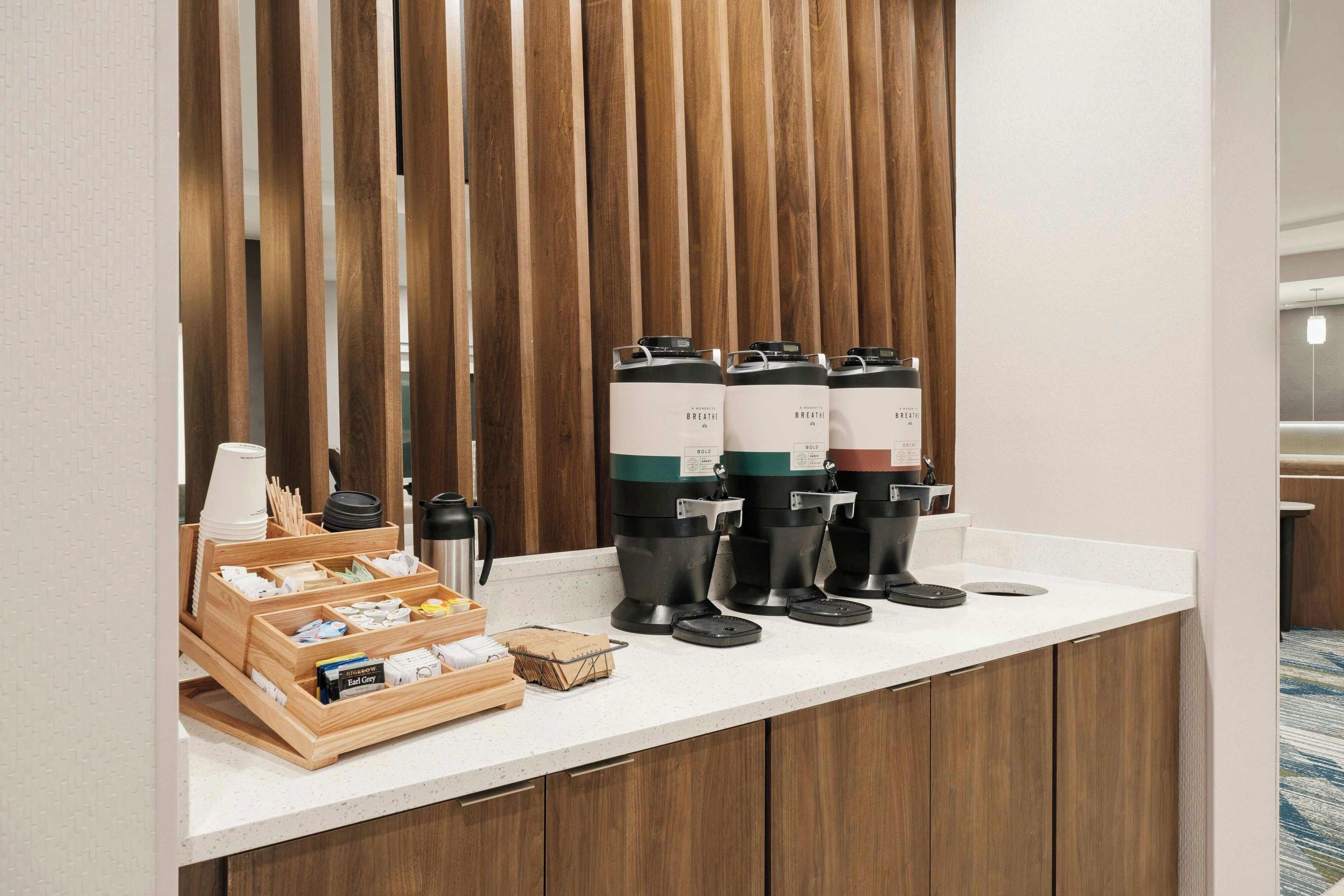 Una estación de café con varios dispensadores, una tetera y una bandeja de vasos desechables.