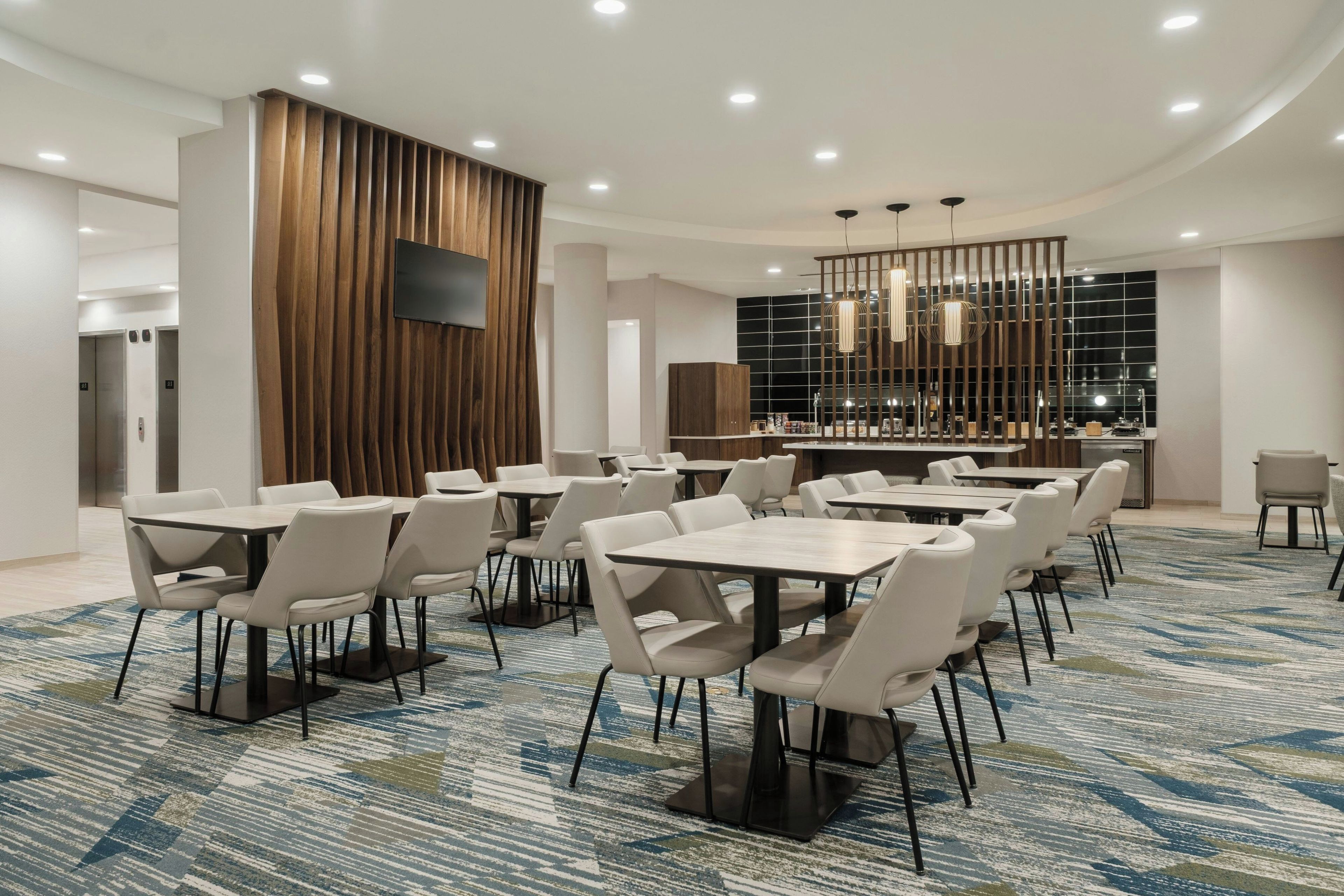 A hotel conference room with multiple tables and chairs, a flat-screen TV mounted on a wooden panel, and a modern chandelier.