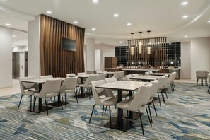 A hotel conference room with multiple tables and chairs, a flat-screen TV mounted on a wooden panel, and a modern chandelier.