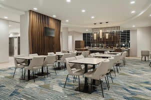 Un salón de conferencias de hotel con múltiples mesas y sillas, un televisor de pantalla plana montado en un panel de madera y una lámpara de araña moderna.