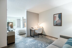 Habitación de hotel con cama, escritorio con silla, mesita pequeña, lámpara y un cuadro en la pared.