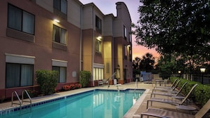 Ein Schwimmbecken mit Liegestühlen und einer Poolterrasse, umgeben von einem Gebäude mit mehreren Fenstern und einer Tür.