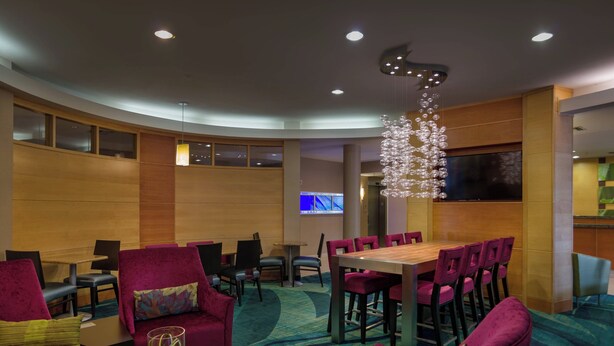 Eine Hotellobby mit einer gebogenen Sitzecke, einem hölzernen Empfangstresen und einem großen Bildschirm, der eine Präsentation zeigt.