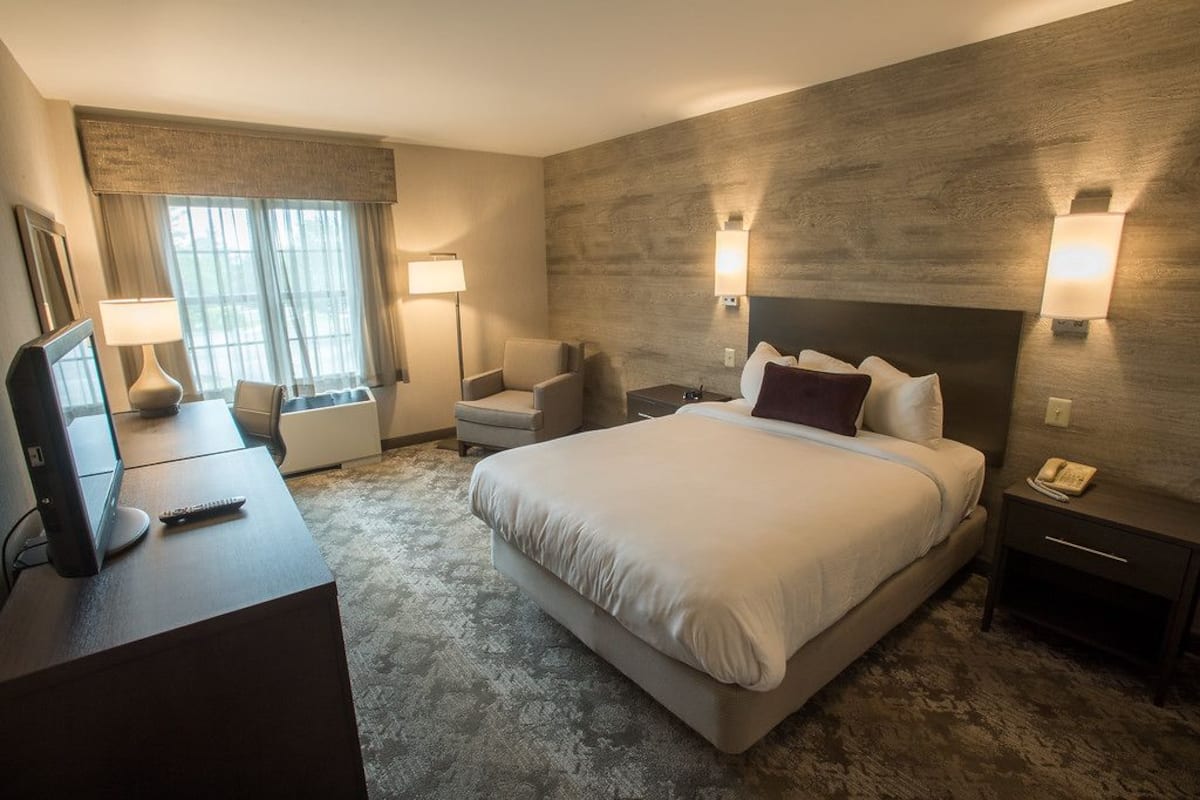 A hotel room with a large bed, a desk with a computer, a chair, and a bedside table with a phone.