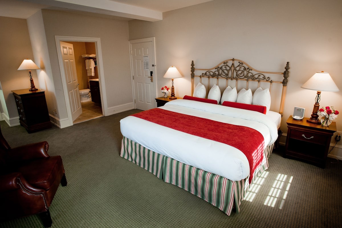 A hotel room with a large bed, two bedside tables with lamps, a wooden chair, and a door leading to a bathroom.