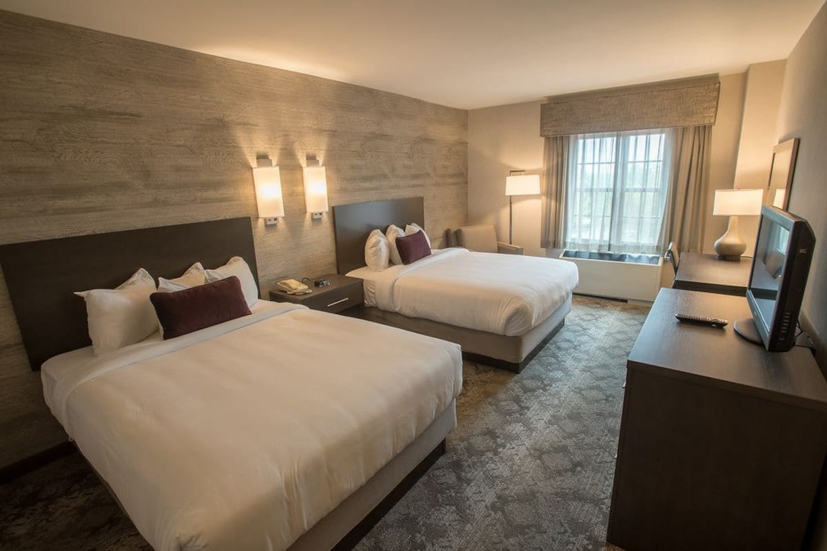 A hotel room with two beds, a desk with a TV, a window with curtains, and wall-mounted lamps.