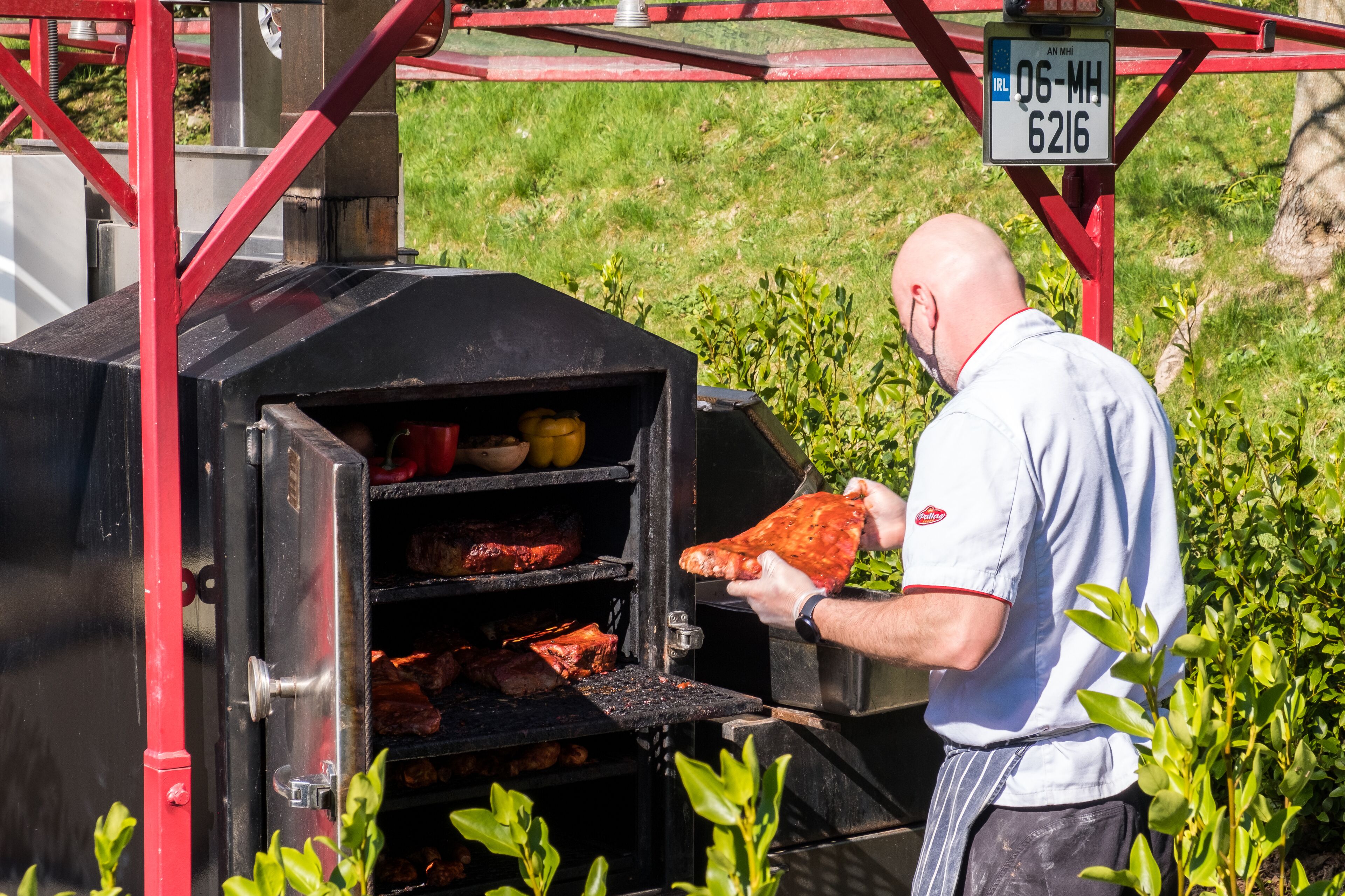 A man grilling meat on an outdoor barbecue with a metal grill and a red frame structure.
