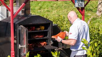 A man grilling meat on an outdoor barbecue with a metal grill and a red frame structure.