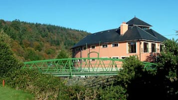 Uma casa de dois andares com telhado escuro e paredes bege, rodeada por vegetação e colinas.