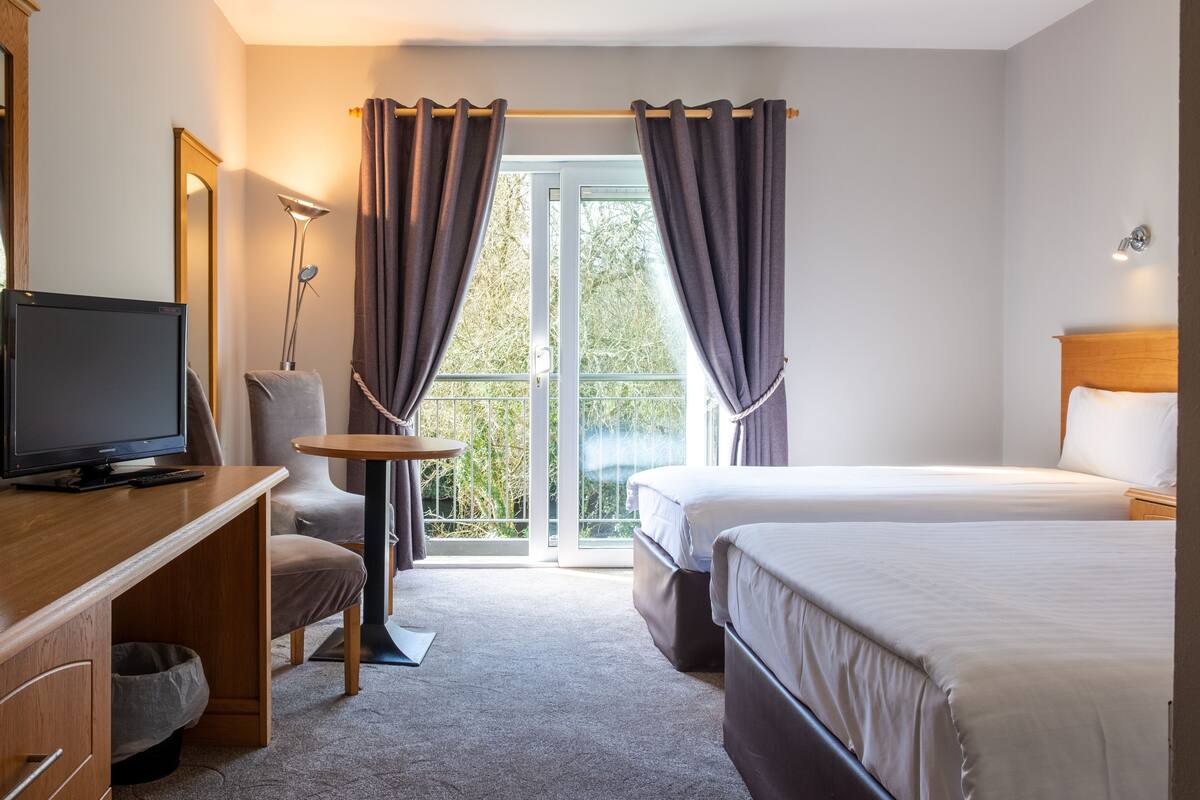 A hotel room with a bed, a desk with a television, a chair, and a balcony with curtains.