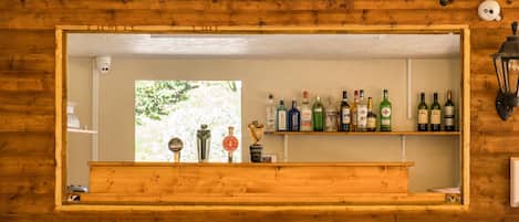 Una zona de bar con barra de madera, estantes llenos de botellas de licor y vistas a zonas verdes al exterior.