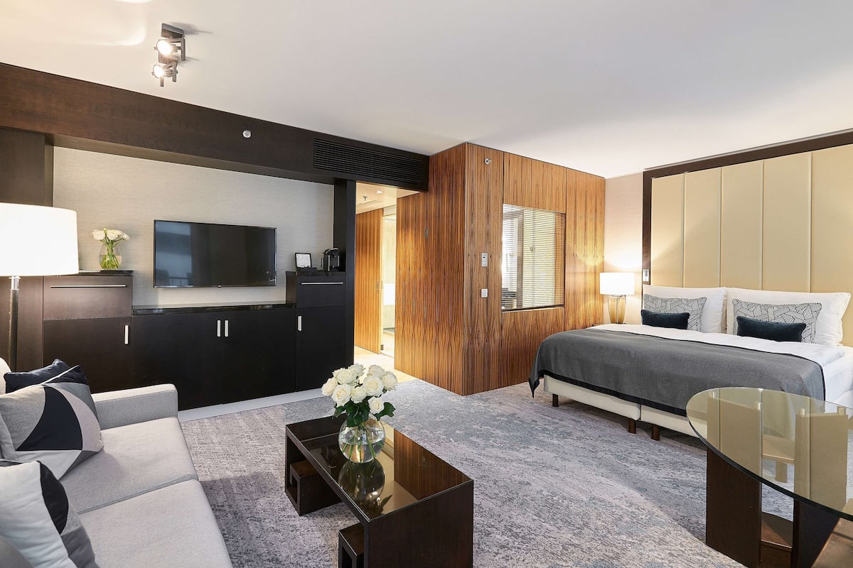 A modern hotel room with a bed, sofa, TV, and wooden paneling.