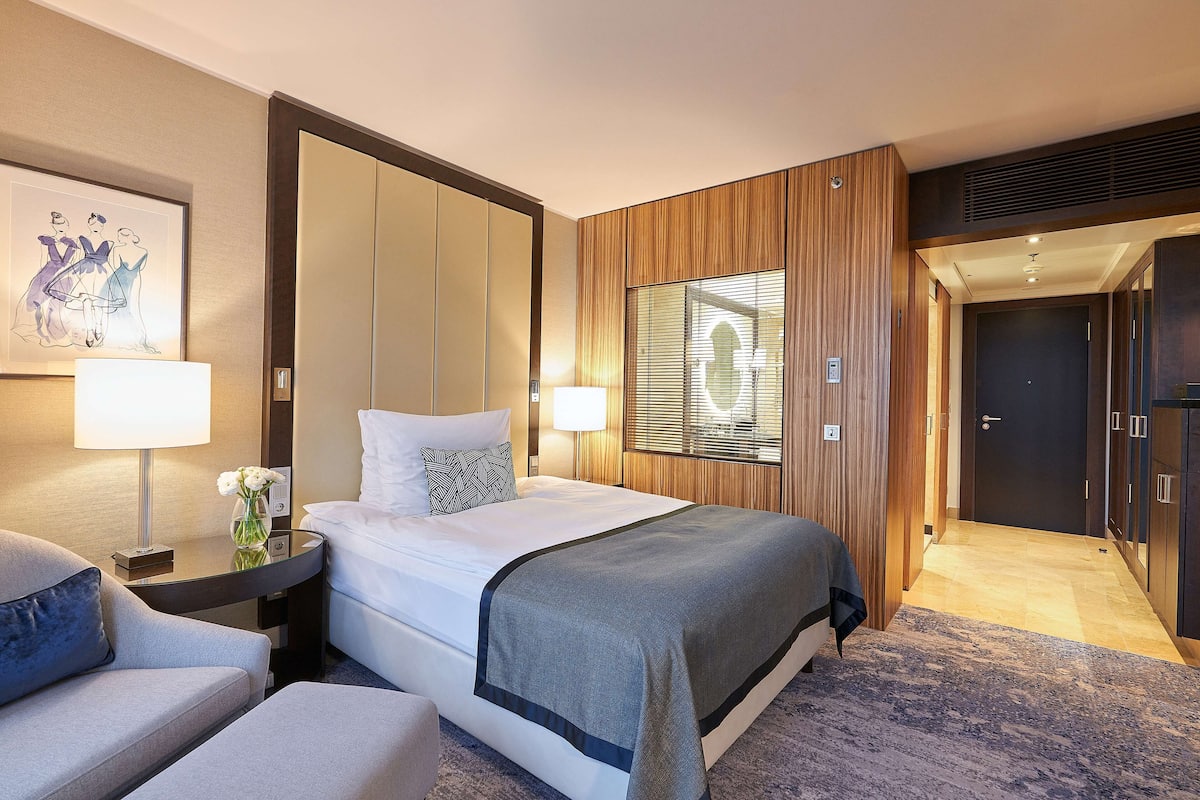 A hotel room with a large bed, a sitting area with a chair, a desk with a lamp, and a framed artwork on the wall.
