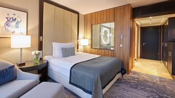 A hotel room with a large bed, a sitting area with a chair, a desk with a lamp, and a framed artwork on the wall.