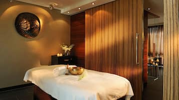 Un spa moderno con una cama, un panel de madera en la pared, un adorno mural y una mesita con vela y flores.