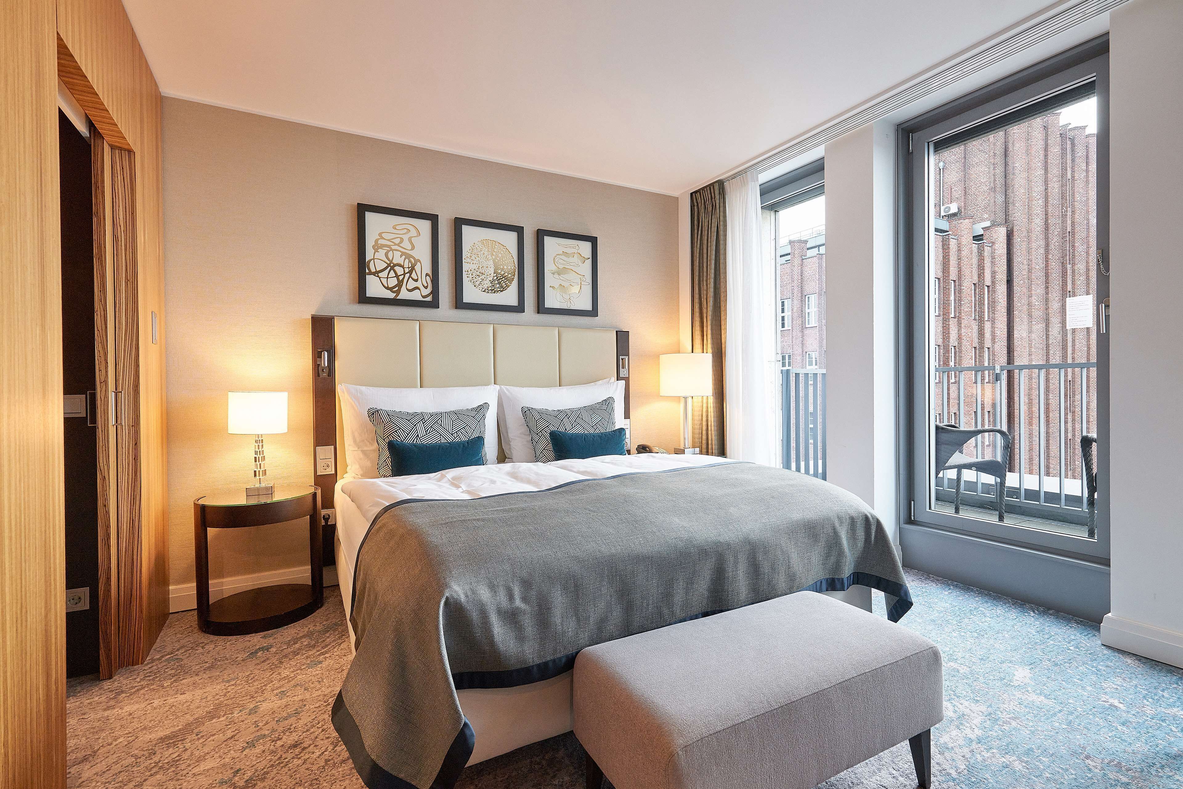 Quarto de hotel com uma cama grande, duas mesas de cabeceira, um banco e vista para um edifício de tijolos através de uma janela ampla.